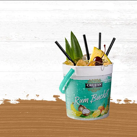 Wide shot of the Rum Runner cocktail in a Cruzan® Rum bucket with drinking straws and garnished with a pineapple wedge and cherry set against a whitewashed wood background with a gold paint swash.