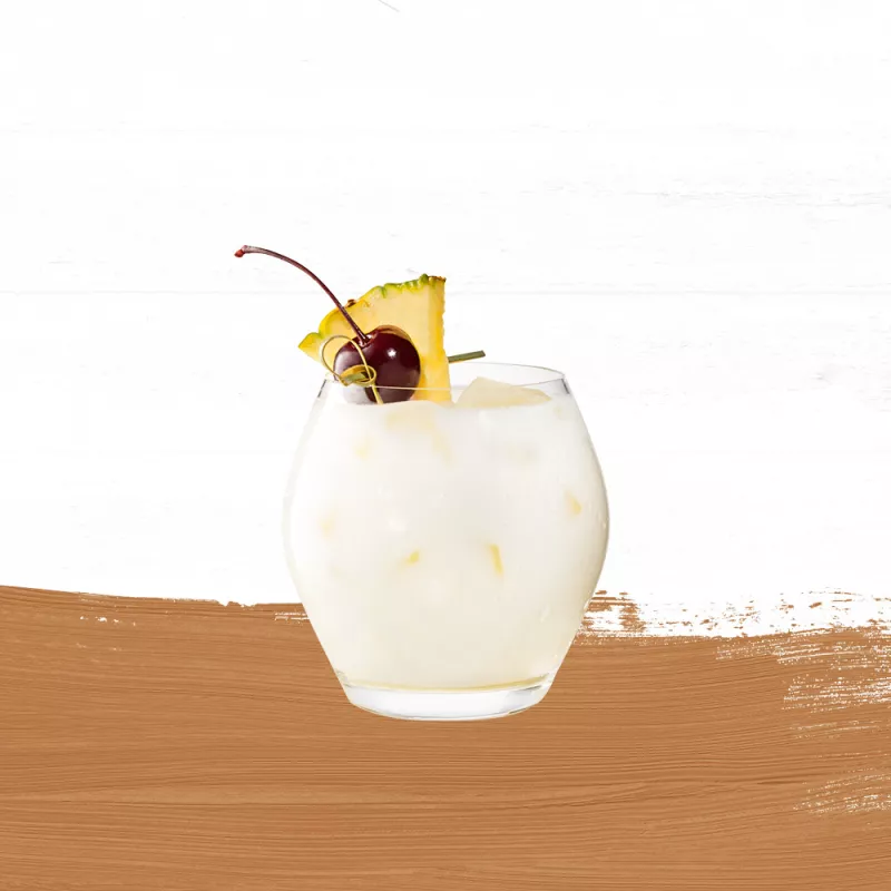 Cruzan® Piña Colada in a clear glass garnished with a cherry and pineapple set against a whitewashed wood background with a gold paint swash.