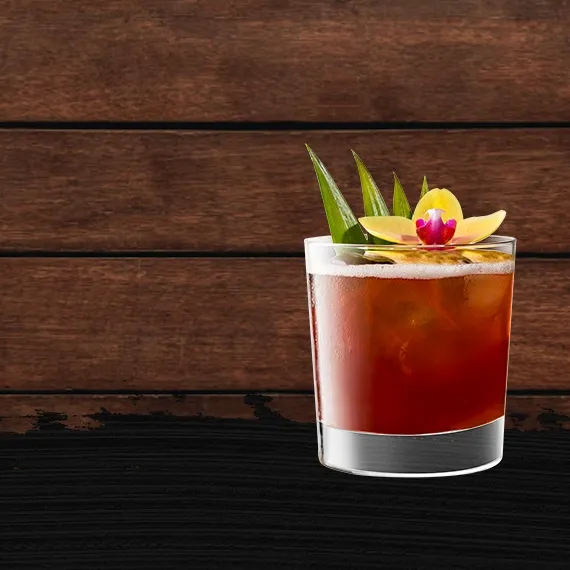 Wide shot of the Cruzan® Jungle Bird cocktail in a double old fashioned glass, garnished with pineapple fronds against a wood background with a black paint swash.